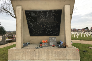 Ein Denkmal auf einem Friedhof mit einer Karte darauf, umgeben von Blumensträußsen und anderen Gegenständen, mit Grabstäten, Bäumen, Gebäuden und einem klaren blauen Himmel im Hintergrund, das das Grab eines im Zweiten Weltkrieg gefallenen deutschen Soldaten markiert.