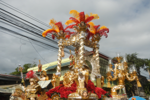 Ein bunt geschmückter Narrentreffen-Wagen mit Statuen und Blumen, umgeben von Gebäuden, Bäumen, Pfählen und Drähten, mit dem Himmel darüber.