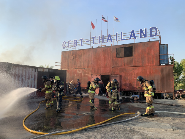 Feuerwehrleute in Helmen sprühen Wasser auf ein Gebäude mit Text, während Flaggen, Bäume und ein blauer Himmel im Hintergrund zu sehen sind.