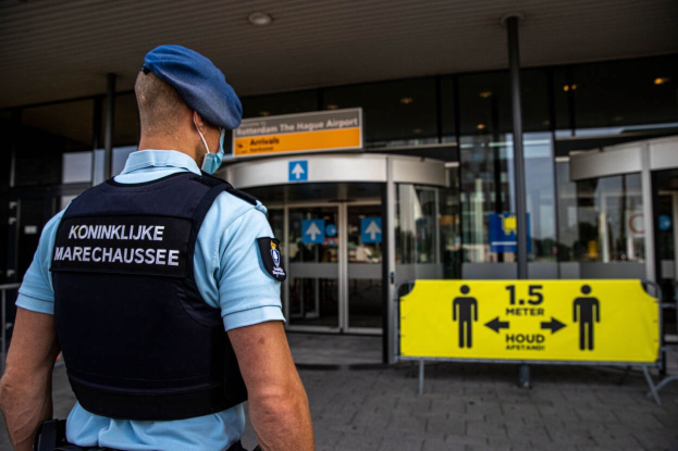 Polizist in blauer Uniform und Maske vor einem Glasgebäude mit gelben und weißen Informationsschildern im Hintergrund.