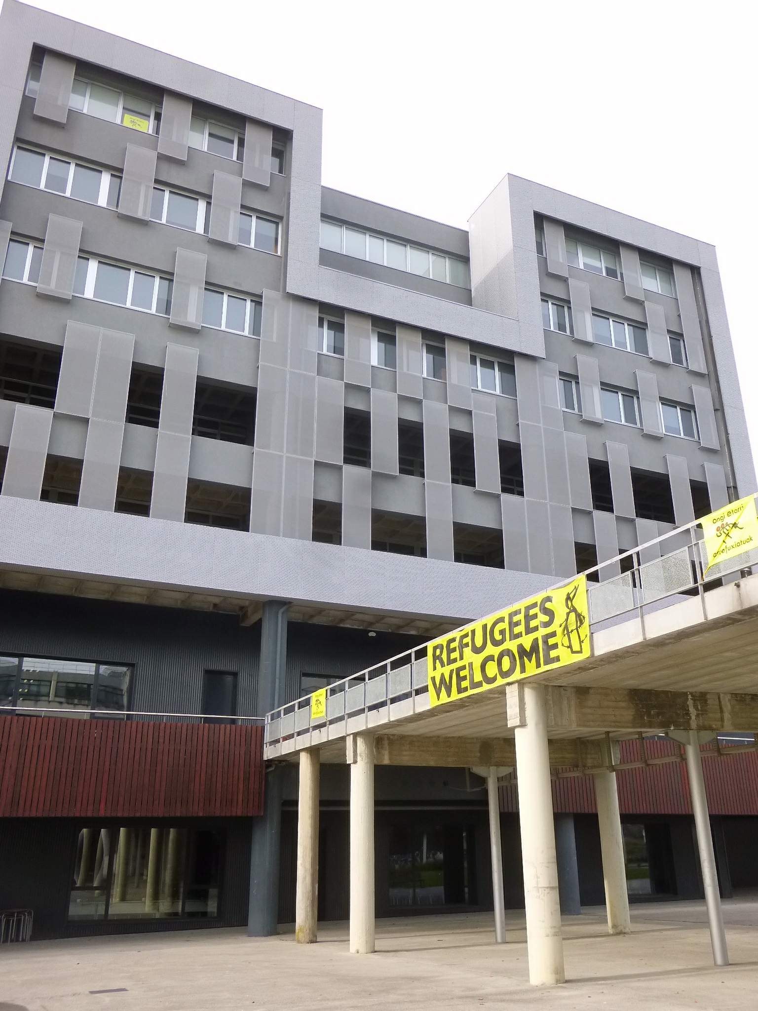 Ein großes Gebäude mit einem "Willkommen bei den Flüchtlingen"-Schild, Säulen, einem Geländer und dem Himmel im Hintergrund.