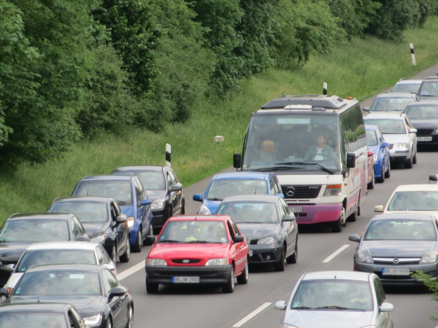Ein Stau auf einer Autobahn mit zahlreichen Autos und einem Lieferwagen, mit Menschen in den Fahrzeugen, mit Bäumen und Gras im Hintergrund.