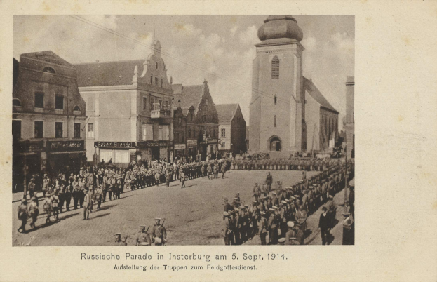Schwarzes und weißes Foto einer Parade in Insterburg im Jahr 1914 mit marschierenden und stehenden Menschen, Gebäuden im Hintergrund und Text unten.