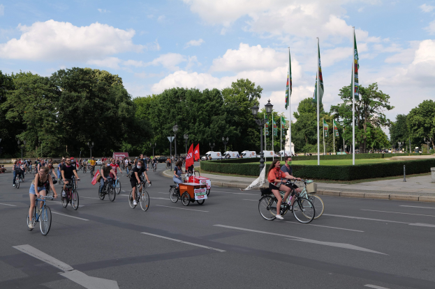 Eine Gruppe von Menschen, die auf Fahrrädern eine von Bäumen gesäumte Straße mit Laternen- und Fahnenmasten entlangfahren, während sie an der Pariser Fahrradtour 2016 teilnehmen.