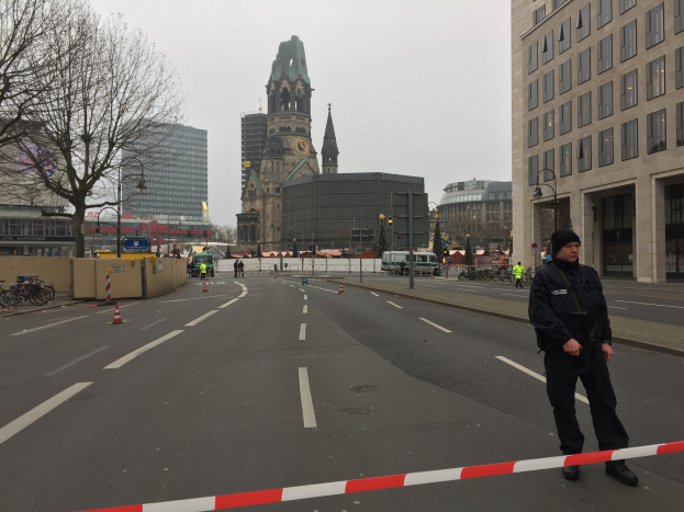 Ein Polizeibeamter in schwarzer Uniform und Mütze steht in einer Berliner Straße umgeben von Verkehrskegeln, Fahrrädern, Laternenmasten, Bäumen und Gebäuden, im Hintergrund ist ein Uhrenturm und ein klarer blauer Himmel zu sehen.