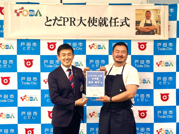 Zwei Männer mit einem Zertifikat in der Hand, das den Text 'Toda City Wins PR Award' trägt, vor einem Foto im Hintergrund.