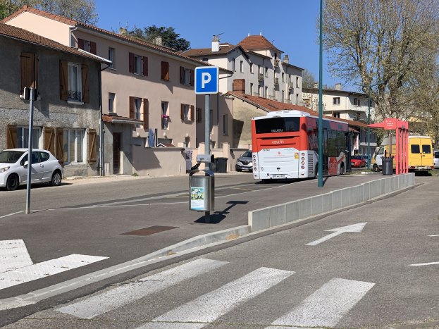 Ein Bus hält an einer Bushaltestelle auf einer Straße mit Gebäuden, Bäumen und Fahrzeugen unter einem klaren blauen Himmel, mit nahegelegenen Wegweisern.