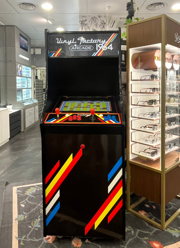 Ein Geschäft mit einem zentralen Vintage-Arcade-Automaten, einem rechten Gestell mit Gläsern und Hintergrund-Elementen wie Schränken, einem Fernseher und anderen Gegenständen, mit Deckenlampen, die eine Retro-Atmosphäre schaffen.