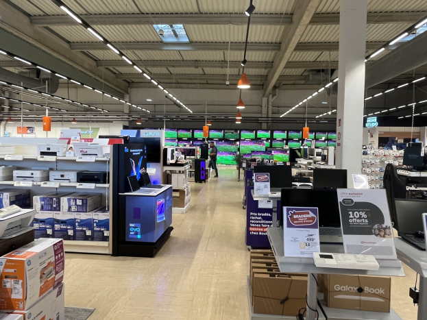 Ein großer Raum in einem Walmart-Laden voller Elektronik wie Computern und Druckern, die in Regalen angeordnet sind, mit Menschen auf dem Boden, einer Säule, Schildern, Deckenleuchten und Ventilatoren.