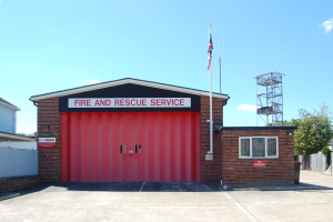 Feuerwehr- und Rettungsdienstgebäude mit roter Tür, Fenstern, einem Namensschild, einer Flagge an einer Flagge, einem Metallturm, einem Zaun, einer Gruppe von Bäumen und einem bewölkten Himmel.