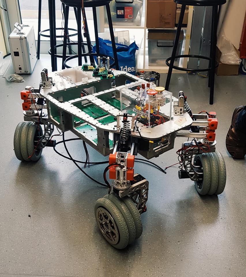 Ein Roboter sitzt auf einem Tisch in einem Raum mit Hockern, Kartons und einem sichtbaren Bein auf der rechten Seite.