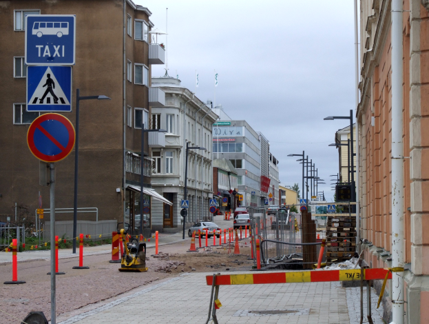Stadtstraßenszene mit Gebäuden, Fahrzeugen, Verkehrsschildern, Baustelle und Bäumen unter einem bewölkten Himmel.