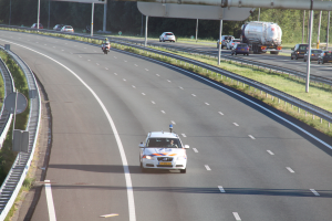 Ein Auto fährt auf einer Autobahn mit einem Polizeiwagen am Straßenrand, anderen Fahrzeugen, Leitplanken, Pfählen, Brettern und Bäumen im Hintergrund.