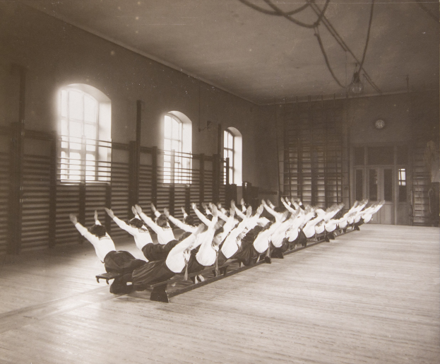 Gruppe von Menschen, die Pilates in einem Fitnessstudio mit Fenstern und einer Uhr an der Wand dahinter machen.