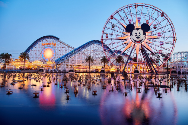 Eine lebendige Dämmerungsszene im Disneyland California Adventure mit einem riesigen Mickey-Maus-Rad im Vordergrund, einer Achterbahn im Hintergrund und Booten auf dem Wasser, mit einem bunten Himmel und silhouettenartigen Bäumen.