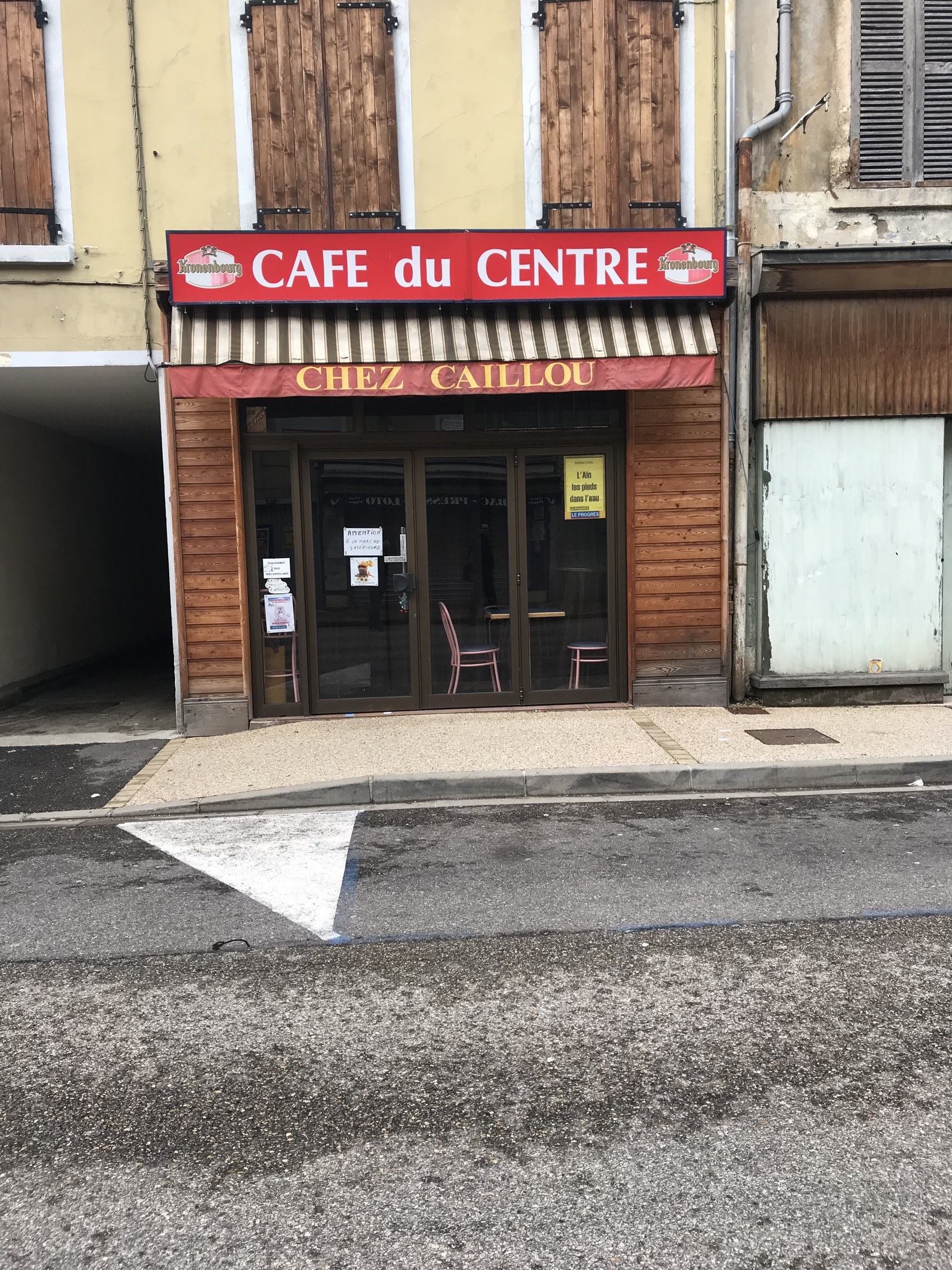 Außenansicht eines Gebäudes mit einer "Cafe du Centre"-Tafel, einer Straße davor und einer Tafel mit Text, mit Fenstern und einer Tür.