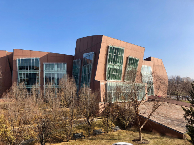 Großes modernes Gebäude mit zahlreichen Fenstern, umgeben von Bäumen, Bänken, Gras und einem Laternenmast, mit anderen Gebäuden und einem klaren blauen Himmel im Hintergrund, als der neue Campus der University of Illinois at Chicago identifiziert.