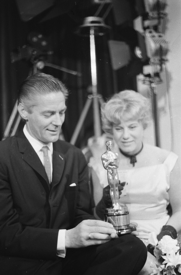 Ein Schwarz-Weiß- Bild eines formell gekleideten Mannes mit einer Oscar-Statue und einer Frau mit einem Blumenstraß in den Händen, die nebeneinander sitzen und von Hintergrundbeleuchtung umgeben sind.