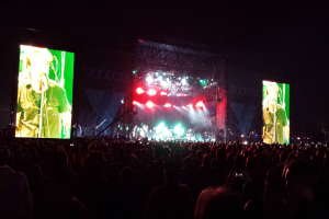 Eine Konzertszene mit einer Menge unten, einem großen Bildschirm links und einem weiteren rechts.