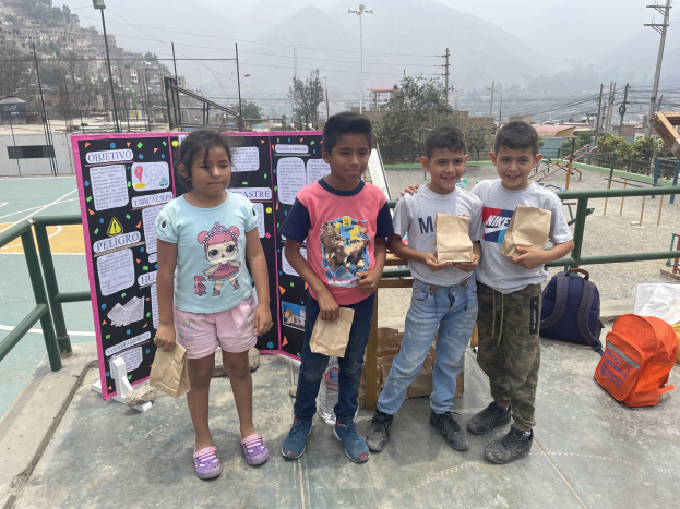 Eine Gruppe von Kindern steht in einer Reihe auf einem Basketballfeld, jeder hält einen Papiertüte in der Hand, mit zusätzlichen Tüten und Gegenständen rechts daneben und einer Tafel mit Text links daneben.