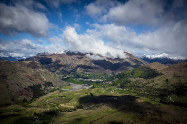 Ein Panoramablick von der Spitze eines Berges in Queenstown, Neuseeland, der saftiges grünes Gras, Bäume und eine gewundene Straße zeigt, unter einem Himmel voller weißer, flauschiger Wolken.