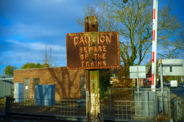 Vorsichtsschild auf einem Bahngleis, umgeben von einem Metallzaun, Bäumen, Strommasten, einem Gebäude, Containern, Fahrzeugen auf einer Straße und einem bewölkten Himmel.