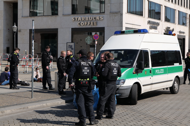 Eine Gruppe von Polizeibeamten steht vor einem Starbucks-Café, rechts davon ein Van und links ein paar Personen; im Hintergrund sind ein Gebäude, ein Schild, ein Laternenpfahl und ein Zaun zu sehen.