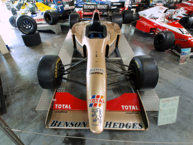 Rennwagen hinter einem Metallzaun in einem Motorsportmuseum, mit einer Texttafel und Hintergrundbesuchern.