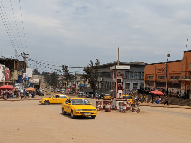 Ein gelbes Taxi fährt auf einer belebten Stadtstraße mit Gebäuden an den Seiten, während Fußgänger mit Schirmen auf dem Gehweg gehen und über Kopfstromleitungen und Wolken am Himmel.