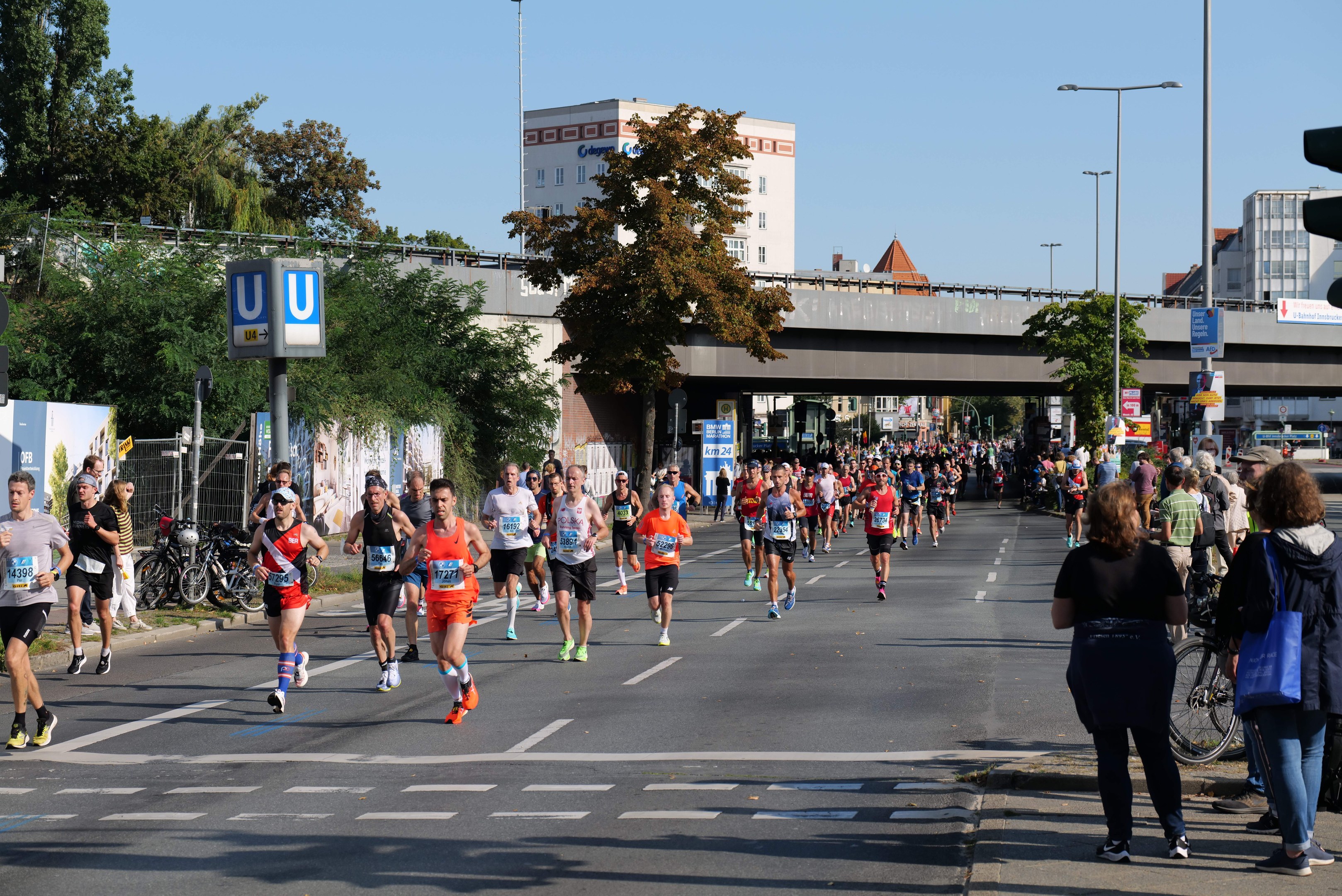 Gruppe von Menschen, die bei einem Marathon auf einer von Bäumen gesäumten Straße mit Laternenpfählen, Schildern, Fahrrädern, einem Zaun, Gras, einer Brücke, Gebäuden und einem klaren blauen Himmel laufen.