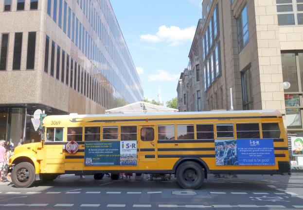Ein gelber Schulbus fährt auf einer Straße mit hohen Gebäuden, Passanten auf dem linken Gehweg, Bäumen im Hintergrund und einem klaren blauen Himmel.