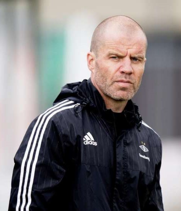 Ein kahlköpfiger Trainer in einer blauen Jacke mit weißen Streifen und weißem Kragen steht auf einem Fußballfeld mit verschränkten Händen und ernster Miene.