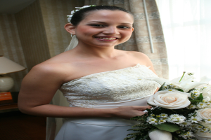 Eine Braut steht strahlend in der Mitte, hält einen Blumenstrauß, mit einem Vorhang und einer Wand im Hintergrund und einer Lampe auf einem Tisch links.