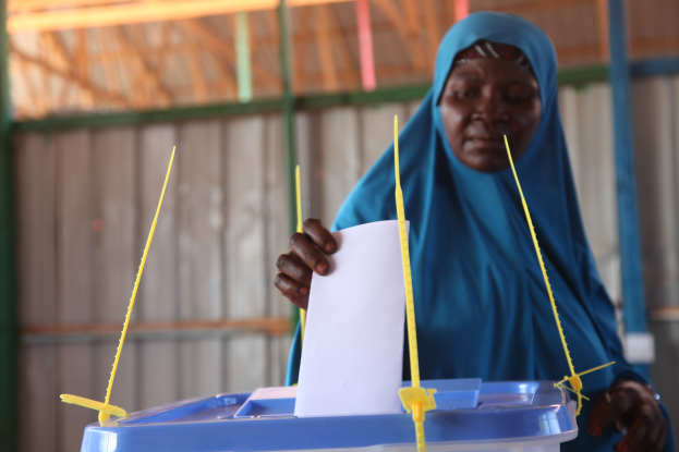 Frau mit blauem Kopftuch, die ein Blatt Papier an einer Wahllokal-Umgebung hält, mit einer Wahlurne und Stöcken davor.
