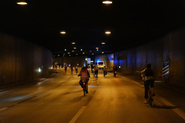 Gruppe von Menschen, die nachts auf Fahrrädern eine Straße entlangfahren, beleuchtet von Lampen von oben, mit Wänden auf beiden Seiten und einem Fahrzeug im Hintergrund.
