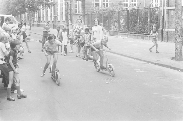Schwarzes und weißes Foto von Kindern, die auf Rollern die Straße hinunterfahren, mit einem geparkten Auto auf der linken Seite, Gebäuden, Bäumen, Pfählen und einem Zaun im Hintergrund.
