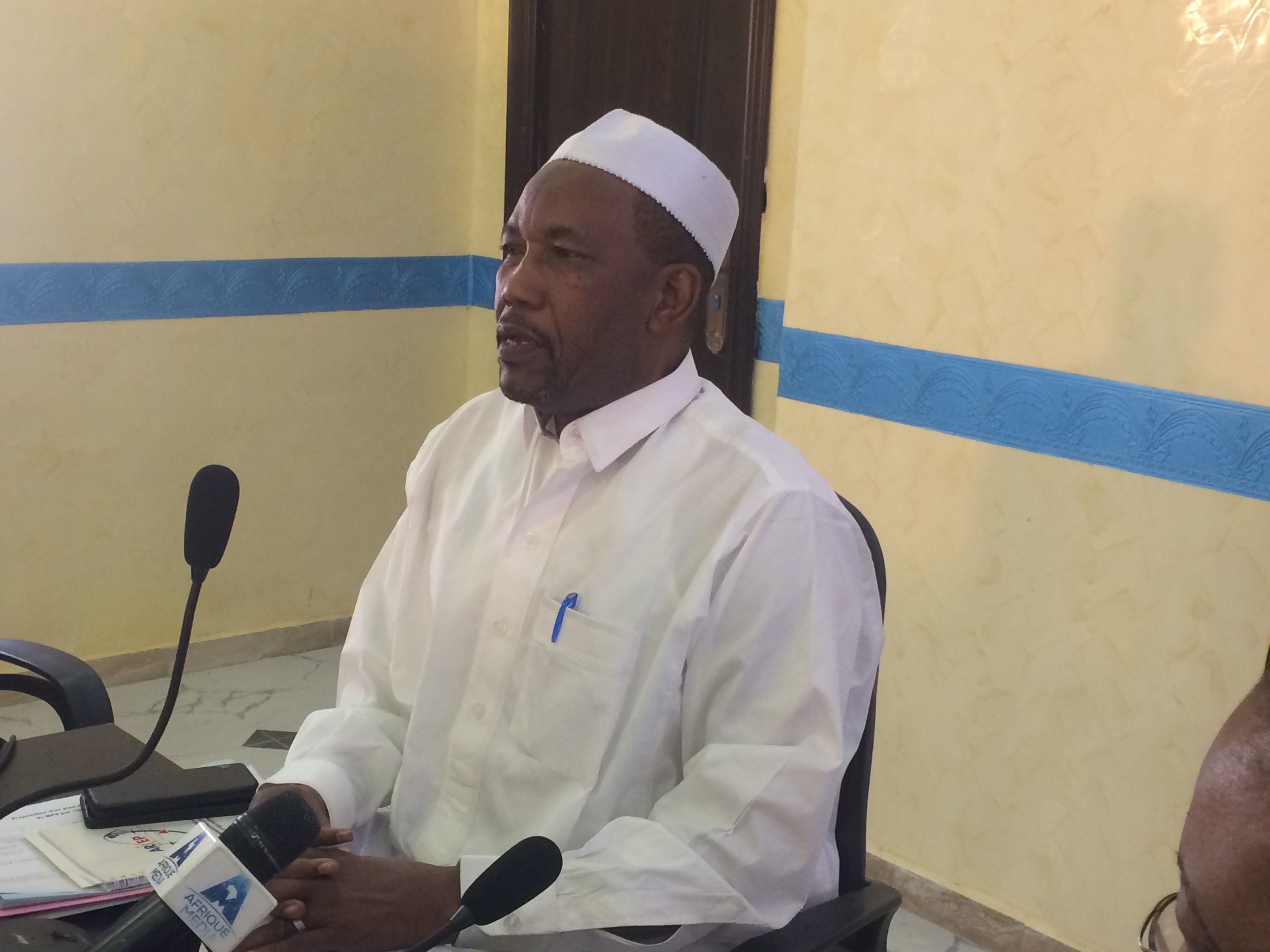 Ein Mann in einem weißen Gewand und Hut sitzt an einem Tisch mit Mikrofonen, während er spricht und Papiere und Gegenstände auf dem Tisch zu sehen sind, mit einer anderen Person mit Brille rechts von ihm, in einem Raum mit einer Wand und einer Tür.