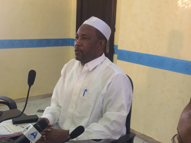 Ein Mann in einem weißen Gewand und Hut sitzt an einem Tisch mit Mikrofonen, während er spricht und Papiere und Gegenstände auf dem Tisch zu sehen sind, mit einer anderen Person mit Brille rechts von ihm, in einem Raum mit einer Wand und einer Tür.