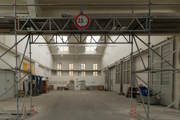 Lagerhalle mit Gerütständen, Verkehrskegeln, Metallstäben, einem Schild, Maschinen, einer Tür, Fenstern, Wänden, Deckenleuchten und einem Dach.