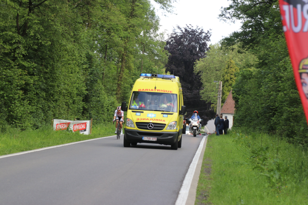 Ambulanz fährt auf einer Straße mit Fahrradfahrern daneben, Gras und Bäume auf beiden Seiten, Häuser, Pfosten und einen klaren blauen Himmel im Hintergrund.