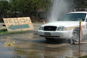 Ein Polizeiwagen fährt durch eine Pfütze, mit einem Schild, einer Verkehrskegel und anderen Gegenständen auf dem Boden, mit Bäumen, Gebäuden und einem klaren blauen Himmel im Hintergrund.