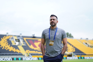 Ein als Ks1 identifizierter Mann, der Trainer der türkischen Nationalmannschaft, steht auf einem Fußballfeld in einem grauen T-Shirt und blauen Hosen, mit gelben Stühlen, einem Baum und einem klaren blauen Himmel im Hintergrund.