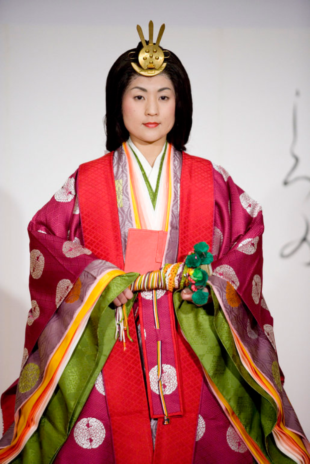 Eine Frau in einem traditionellen japanischen Kimono, das ein leuchtend rotes und grünes Kleid mit aufwändigen Mustern trägt, steht selbstbewusst auf einem Laufsteg, ihr Haar ist ordentlich zurückgebunden.