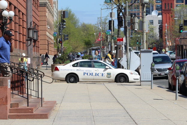 Eine belebte Stadtstraße mit einem parkenden Polizeiwagen, fahrenden Fahrzeugen, Fußgängern, Gebäuden, Laternen, Geländern, Bäumen und einem klaren blauen Himmel.