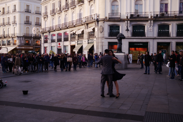 Ein Mann und eine Frau tanzen auf einem belebten Stadtplatz, umgeben von Zuschauern, mit einem Kinderwagen, einem Eimer und Polen links und Gebäuden, Geländern, Schildern, einer Statue und einem bewölkten Himmel im Hintergrund.