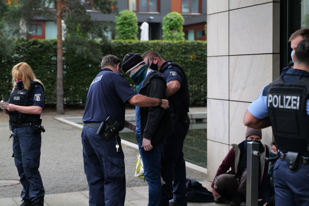 Deutsche Polizeibeamte stehen in einer Gruppe vor einem Gebäude mit einer Frau, die ein Buch und einen Stift hält, während andere in der Nähe sitzen oder stehen, mit Bäumen und Gebäuden im Hintergrund.