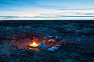 Ein Lagerfeuer mit Menschen, die darum sitzen und liegen, mit Mützen tragend, sowie einem Rucksack und anderen Gegenständen auf dem Boden, umgeben von Bäumen und Himmel.