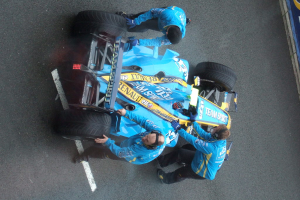 Ein blaues Auto auf einer Straße mit drei Personen, die jeweils am Auto festhalten.