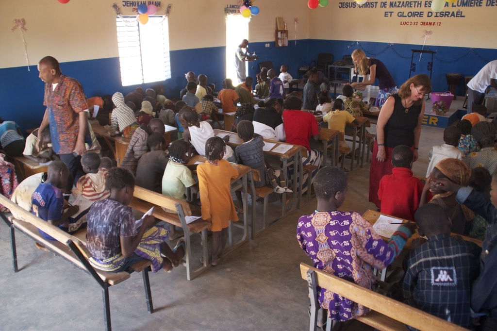Kinder sitzen auf Bänken im Vordergrund, mit Erwachsenen, die sie führen, und einem Hintergrund mit Stühlen, einer Tür, einem Fenster, einem Tisch, Ballons und Text oben.