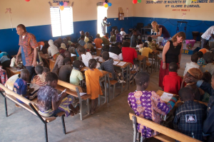 Kinder sitzen auf Bänken im Vordergrund, mit Erwachsenen, die sie führen, und einem Hintergrund mit Stühlen, einer Tür, einem Fenster, einem Tisch, Ballons und Text oben.